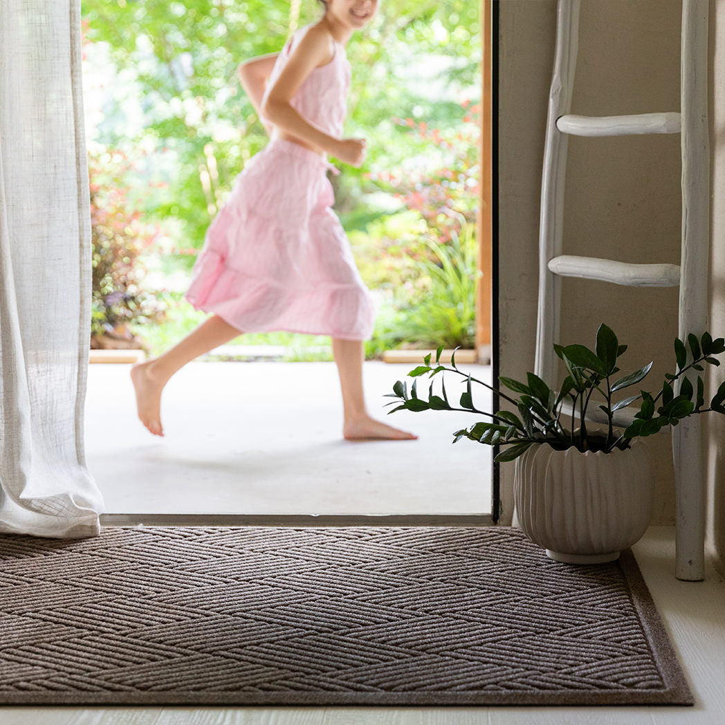 The WaterHog Diamonds Doormat in a camel tan color rests at the base of an open doorway. Its raised, crisscrossing lines form a repeating diamond pattern that adds texture and depth to the entryway. A barefoot child in a pink dress runs past outside, while soft light filters through sheer curtains and a white planter adds a calm, natural touch beside the mat.