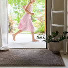 The WaterHog Diamonds Doormat in a camel tan color rests at the base of an open doorway. Its raised, crisscrossing lines form a repeating diamond pattern that adds texture and depth to the entryway. A barefoot child in a pink dress runs past outside, while soft light filters through sheer curtains and a white planter adds a calm, natural touch beside the mat.