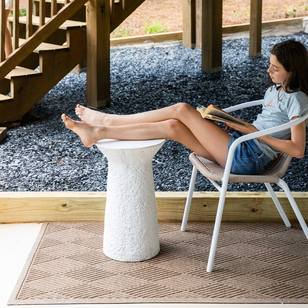 The WaterHog Diamonds Doormat in a camel tan color lies beneath a white accent table and chair, where a young girl relaxes with a book. The mat features a raised diamond pattern made up of angled lines that form a clean, geometric design. Its warm neutral tone contrasts subtly against the pale flooring, bringing a touch of texture and structure to the cozy outdoor reading spot.