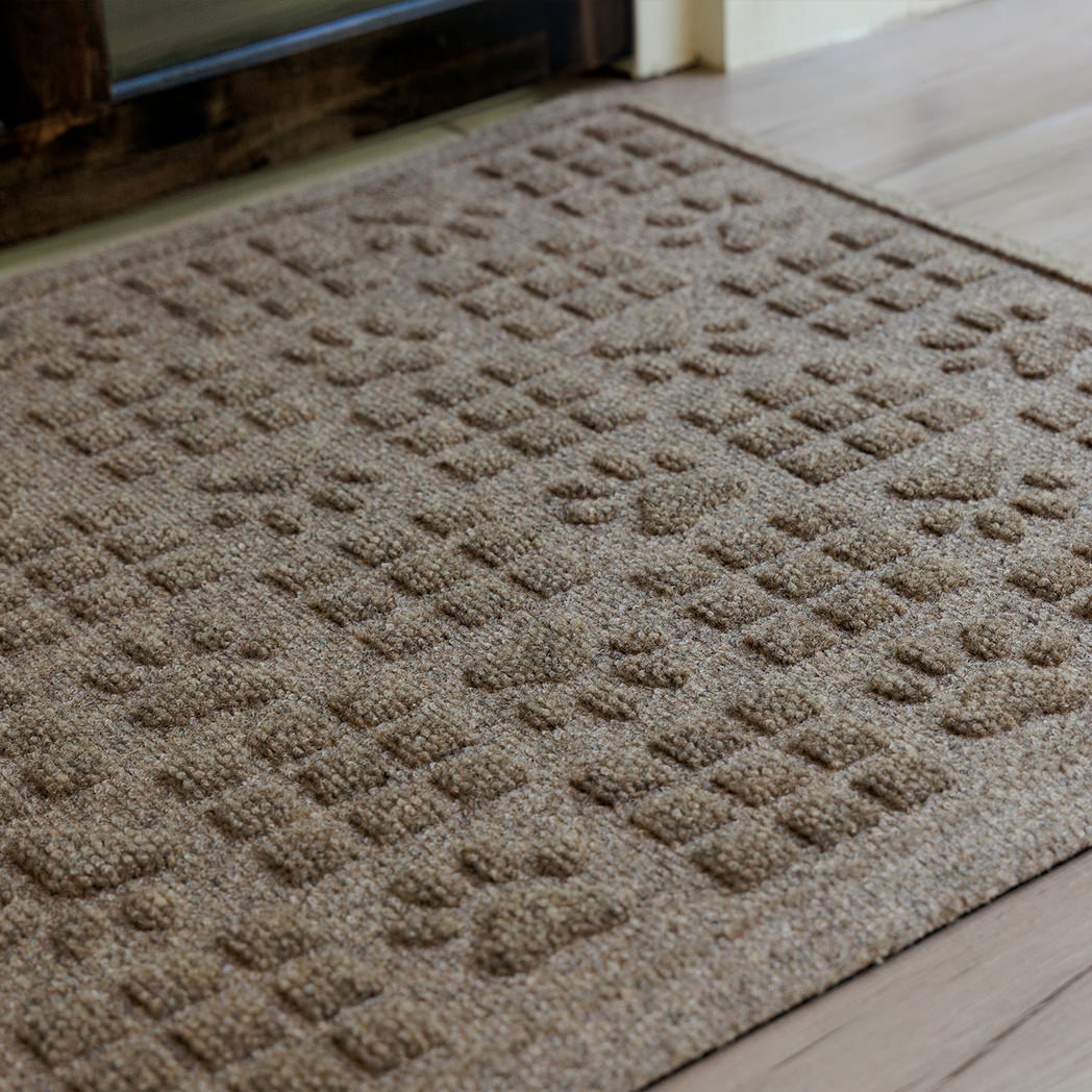 The WaterHog Dog Paw Squares Doormat in beige sits on a pale wood floor near a glass door. Its playful design combines raised paw prints and small square tiles arranged in a repeating pattern across the mat’s surface. The textured weave gives it a cozy, durable look—perfect for homes with pets tracking in a bit of adventure from outside.