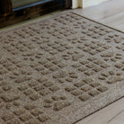 The WaterHog Dog Paw Squares Doormat in beige sits on a pale wood floor near a glass door. Its playful design combines raised paw prints and small square tiles arranged in a repeating pattern across the mat’s surface. The textured weave gives it a cozy, durable look—perfect for homes with pets tracking in a bit of adventure from outside.