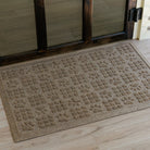 The WaterHog Dog Paw Squares Doormat in a warm tan color sits on a light wood floor by a glass door. Its raised design alternates between clusters of small square tiles and larger pawprints, giving the surface a fun, textured feel. The soft beige tones blend naturally with the wood flooring, creating a cozy, pet-friendly entryway that's both stylish and functional.