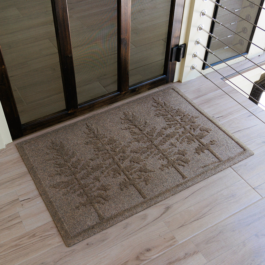The WaterHog Fir Lineup Doormat in a warm tan color sits on a light wood floor near a glass door. Its surface features a row of tall, detailed fir trees sculpted into the fibers, their branches rising in clean vertical symmetry. The textured design adds depth and character, giving the mat a natural, outdoors-inspired look that complements the soft, earthy tones of the entryway.
