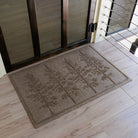 The WaterHog Fir Lineup Doormat in a warm tan color sits on a light wood floor near a glass door. Its surface features a row of tall, detailed fir trees sculpted into the fibers, their branches rising in clean vertical symmetry. The textured design adds depth and character, giving the mat a natural, outdoors-inspired look that complements the soft, earthy tones of the entryway.