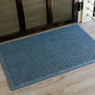 The WaterHog Hampton Doormat in a soft blue-gray color lies on a light wood floor in front of a dark-framed glass door. Its surface displays a uniform grid of raised, pill-shaped ridges that create a subtle, woven brick texture. The overhead angle highlights the mat’s crisp edges and clean alignment, adding a sense of order to the entryway.