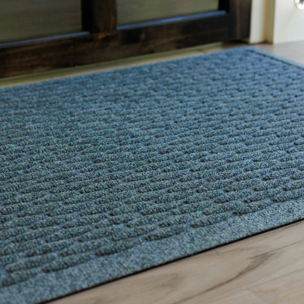 The WaterHog Hampton Doormat in a cool blue-gray tone rests in front of a dark wooden door. Its surface features a neatly arranged pattern of raised rectangular ridges that resemble a cobblestone texture. The mat’s subtle color variation and low profile blend naturally with the light wood flooring, creating a clean, structured entryway look.