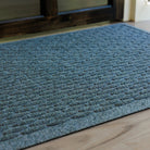 The WaterHog Hampton Doormat in a cool blue-gray tone rests in front of a dark wooden door. Its surface features a neatly arranged pattern of raised rectangular ridges that resemble a cobblestone texture. The mat’s subtle color variation and low profile blend naturally with the light wood flooring, creating a clean, structured entryway look.