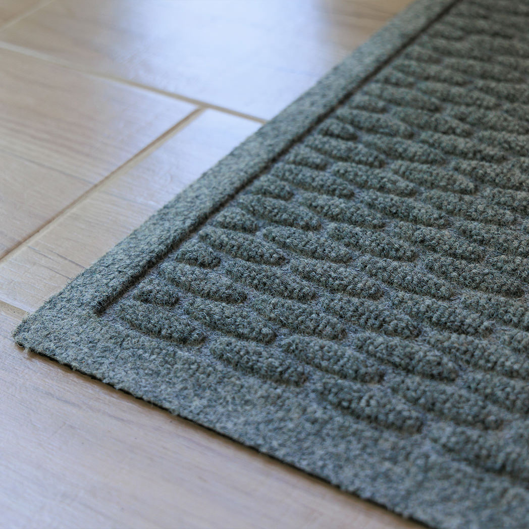 The WaterHog Hampton Doormat in a muted blue-gray color sits on a light wood floor. The mat features a raised brick-like pattern made up of short, rounded rectangular ridges that add subtle texture and depth. The close-up angle highlights the mat’s sturdy fibers and defined edges, emphasizing its durable, structured design.