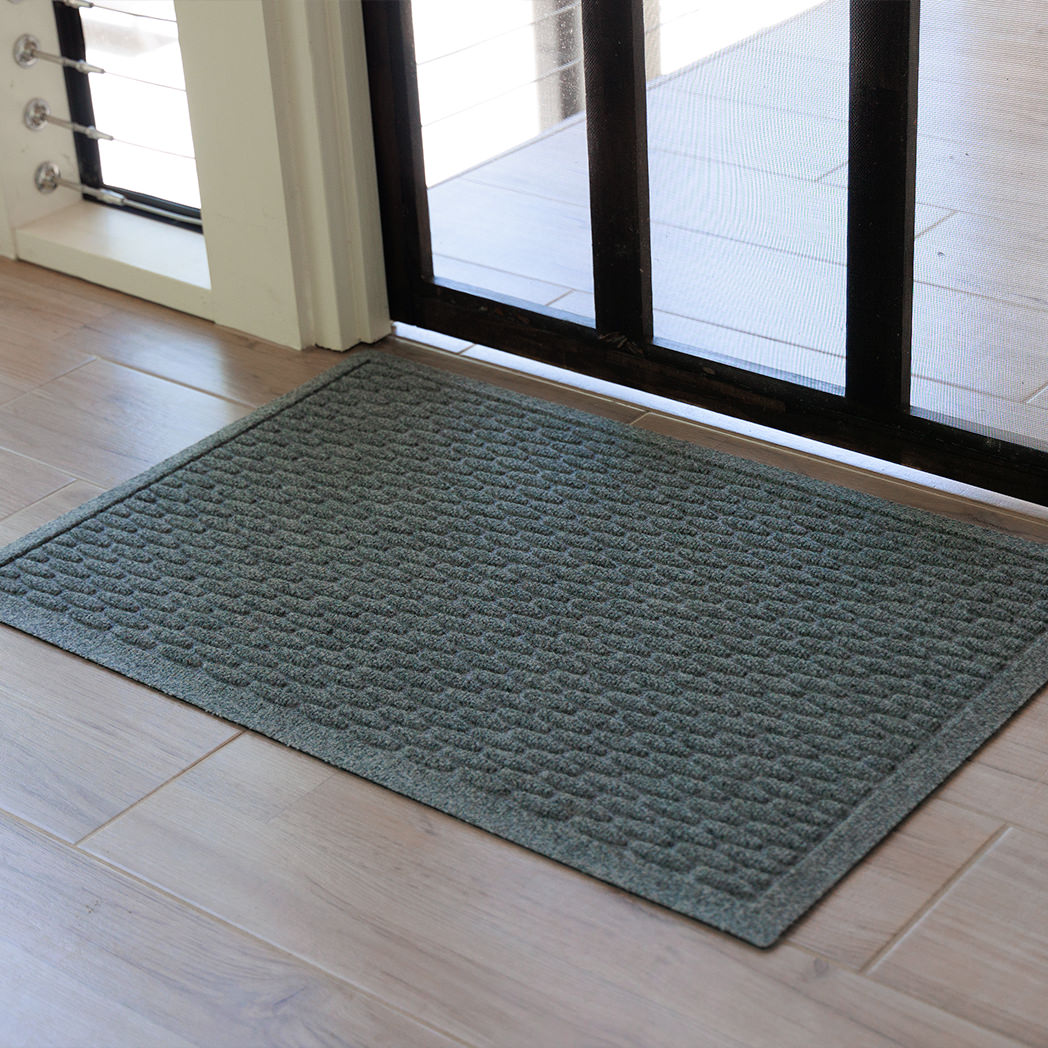 The WaterHog Hampton Doormat in gray rests on a light wood tile floor just inside a glass door with a dark frame. Its surface features an even grid of raised rectangular ridges that create a textured, brick-like appearance. The natural light from the doorway softens the gray tone, highlighting the mat’s clean edges and precise, structured design.