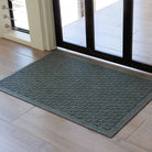 The WaterHog Hampton Doormat in gray rests on a light wood tile floor just inside a glass door with a dark frame. Its surface features an even grid of raised rectangular ridges that create a textured, brick-like appearance. The natural light from the doorway softens the gray tone, highlighting the mat’s clean edges and precise, structured design.