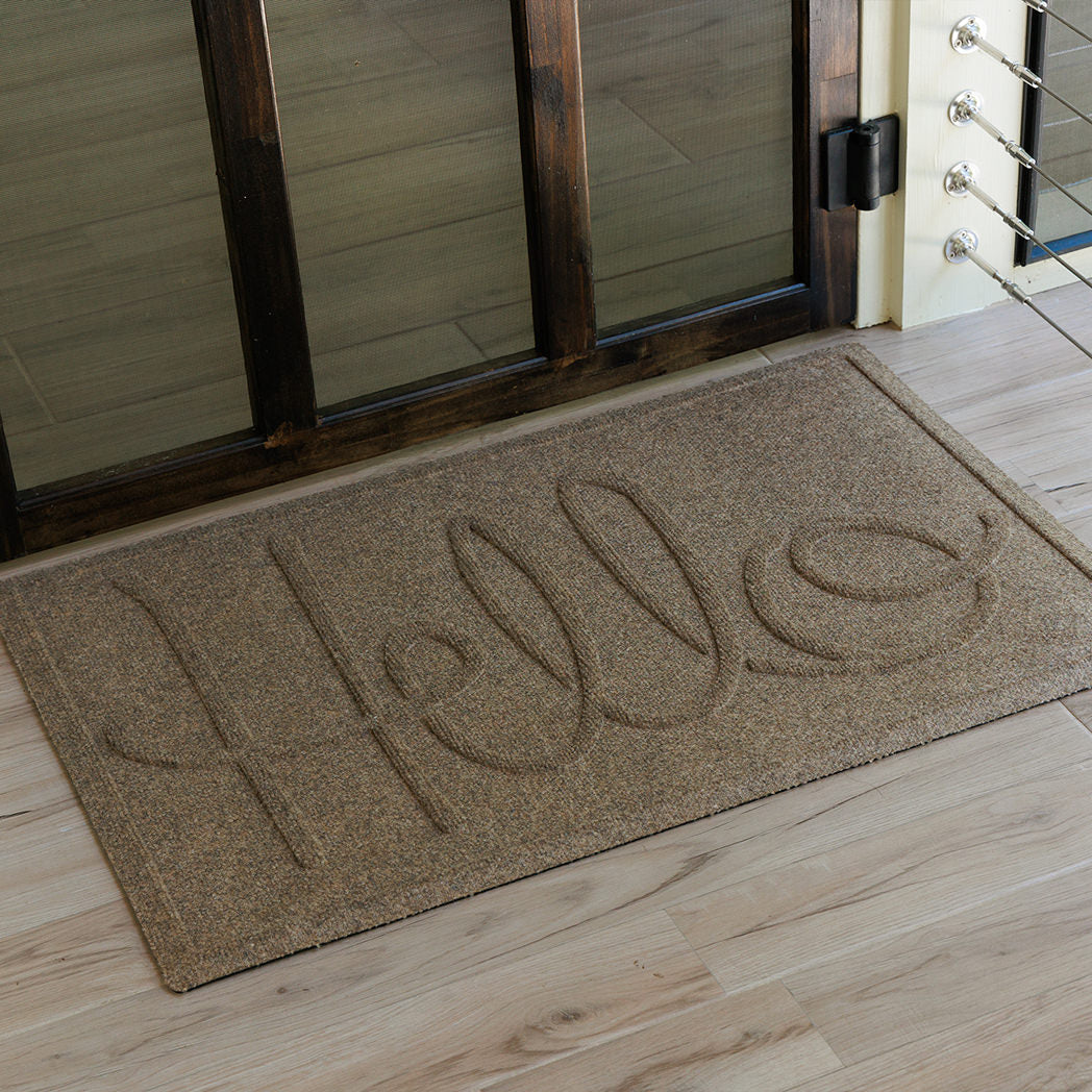 The WaterHog Hello Doormat in a camel tan color rests outside a dark wooden door with glass panels. The mat features the word “Hello” in large, looping raised script across its textured surface. Soft daylight highlights the subtle two-tone fibers of the mat, contrasting against the pale wood decking beneath it.