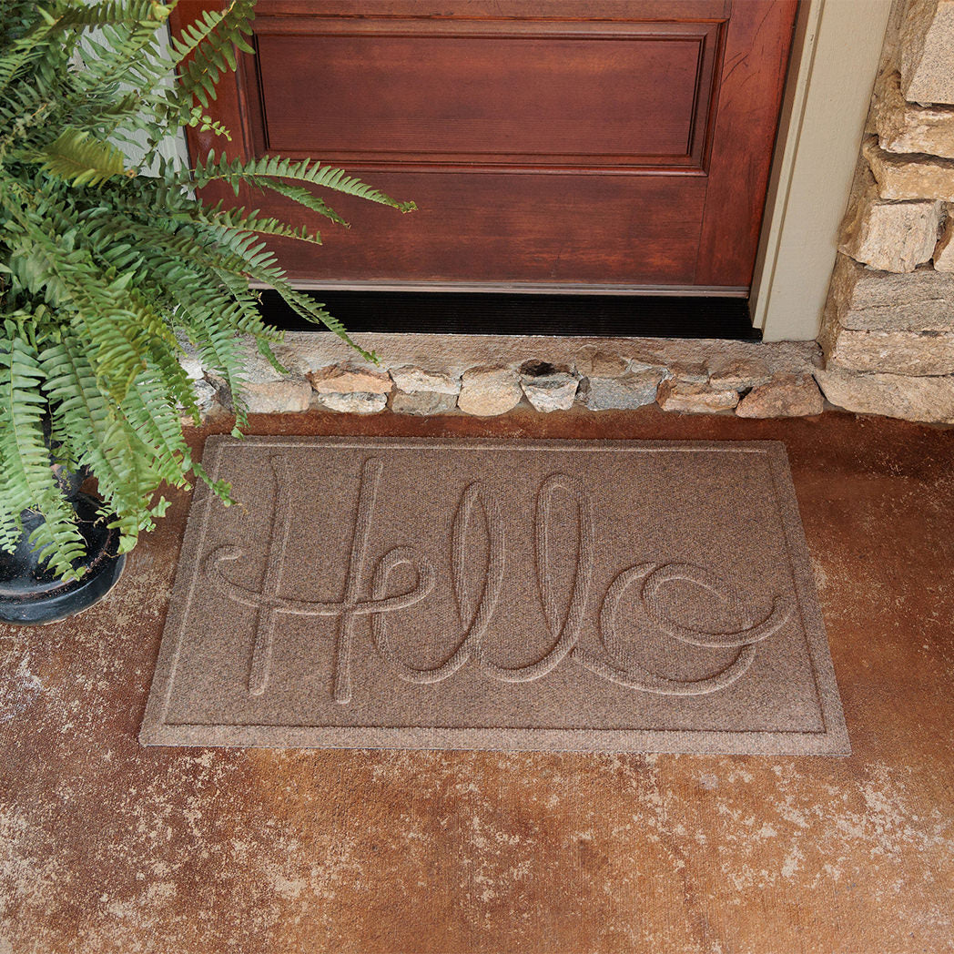 The WaterHog Hello Doormat sits on a dark tan concrete porch in front of a wooden door framed by stone siding. The mat is a warm camel color with the word “Hello” written across the surface in large, looping raised letters. A potted fern adds greenery to the corner, softening the textured stone and concrete around the entryway.