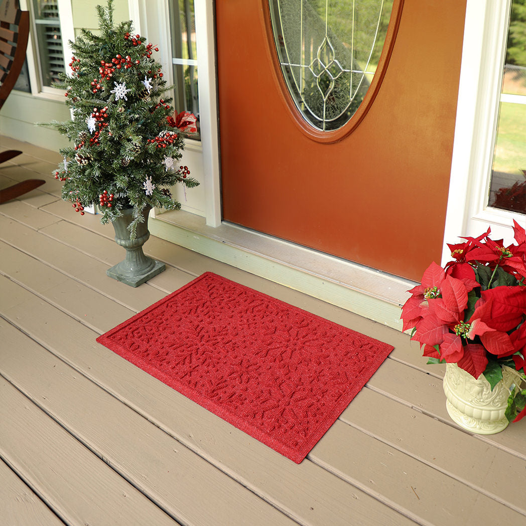 Bright Red holiday snowflake doormat on a wooden deck with a Christmas tree and potted plant.