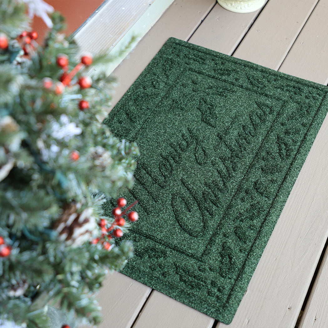 A Deep green, bi-level Merry Christmas doormat on a wooden floor next to a mini Christmas tree.