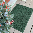 A Deep green, bi-level Merry Christmas doormat on a wooden floor next to a mini Christmas tree.