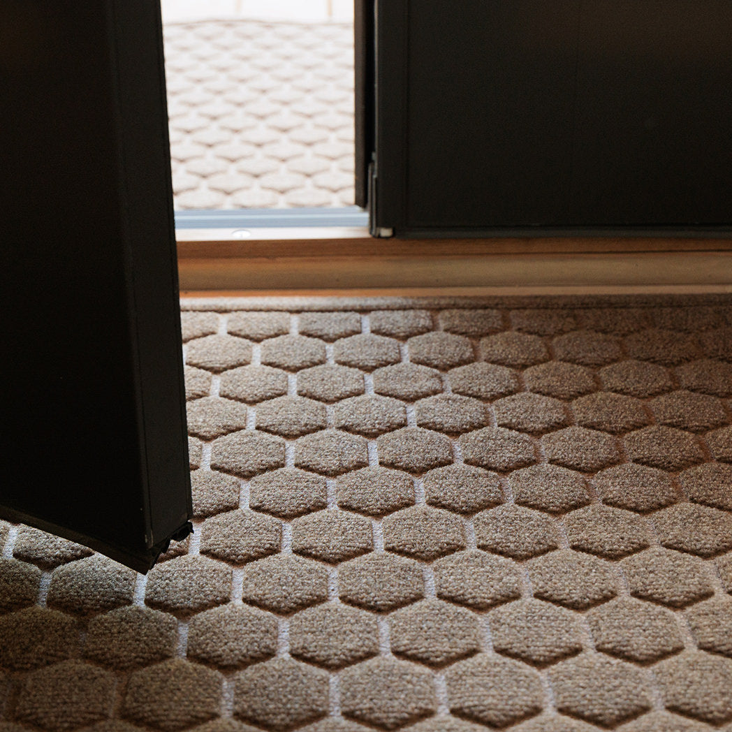 The WaterHog Honeycomb Doormat rests just inside an open doorway, with another matching mat visible outside. Both are a medium beige color, similar to light tan wood, and feature a raised hexagon pattern that forms a honeycomb texture. Sunlight from outside casts soft highlights across the surface, emphasizing the three-dimensional design beneath the open black door.