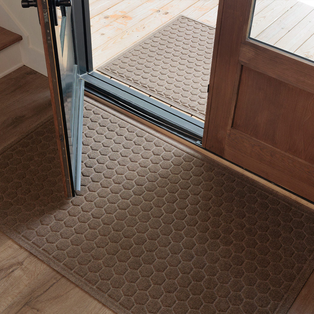 A tan WaterHog Honeycomb Large Doormat rests inside a wooden doorway, with a matching Honeycomb Runner placed on the porch outside. The repeating hexagonal pattern adds a touch of structure and symmetry to the entryway, bridging the transition between indoor floors and the outdoor deck.