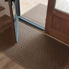 A tan WaterHog Honeycomb Large Doormat rests inside a wooden doorway, with a matching Honeycomb Runner placed on the porch outside. The repeating hexagonal pattern adds a touch of structure and symmetry to the entryway, bridging the transition between indoor floors and the outdoor deck.