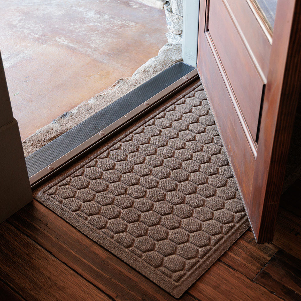 Tan WaterHog Honeycomb Doormat rests just inside an open wooden door on dark hardwood floors. Its pattern of raised hexagons creates a tidy grid that adds texture and structure to the entryway, framed by warm natural light from outside.