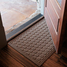 Tan WaterHog Honeycomb Doormat rests just inside an open wooden door on dark hardwood floors. Its pattern of raised hexagons creates a tidy grid that adds texture and structure to the entryway, framed by warm natural light from outside.