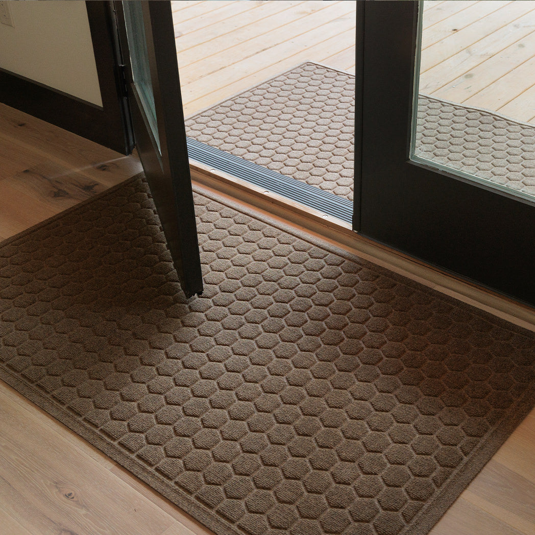A tan WaterHog Honeycomb Large Doormat sits just inside a set of open glass doors, paired with a matching Honeycomb Runner on the deck outside. Both mats feature a raised hexagonal pattern that creates a clean, structured look while helping to trap dirt and moisture between indoor and outdoor spaces.