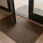 A tan WaterHog Honeycomb Large Doormat sits just inside a set of open glass doors, paired with a matching Honeycomb Runner on the deck outside. Both mats feature a raised hexagonal pattern that creates a clean, structured look while helping to trap dirt and moisture between indoor and outdoor spaces.