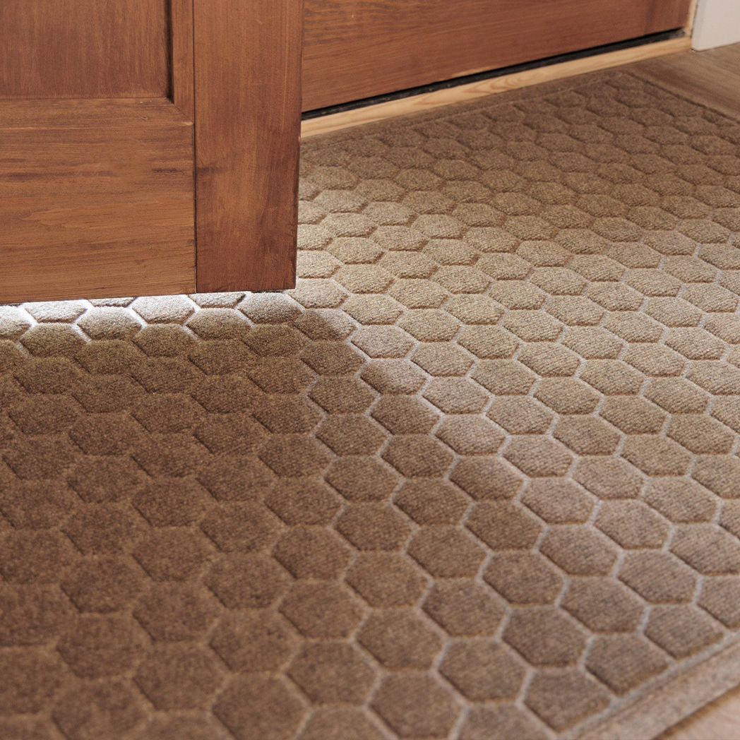 The WaterHog Honeycomb Doormat in a warm tan hue sits just inside a wooden doorway, its hexagonal pattern highlighted by soft natural light. The raised texture adds depth and contrast against the light wood flooring, blending function with a clean, inviting look.