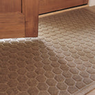 The WaterHog Honeycomb Doormat in a warm tan hue sits just inside a wooden doorway, its hexagonal pattern highlighted by soft natural light. The raised texture adds depth and contrast against the light wood flooring, blending function with a clean, inviting look.