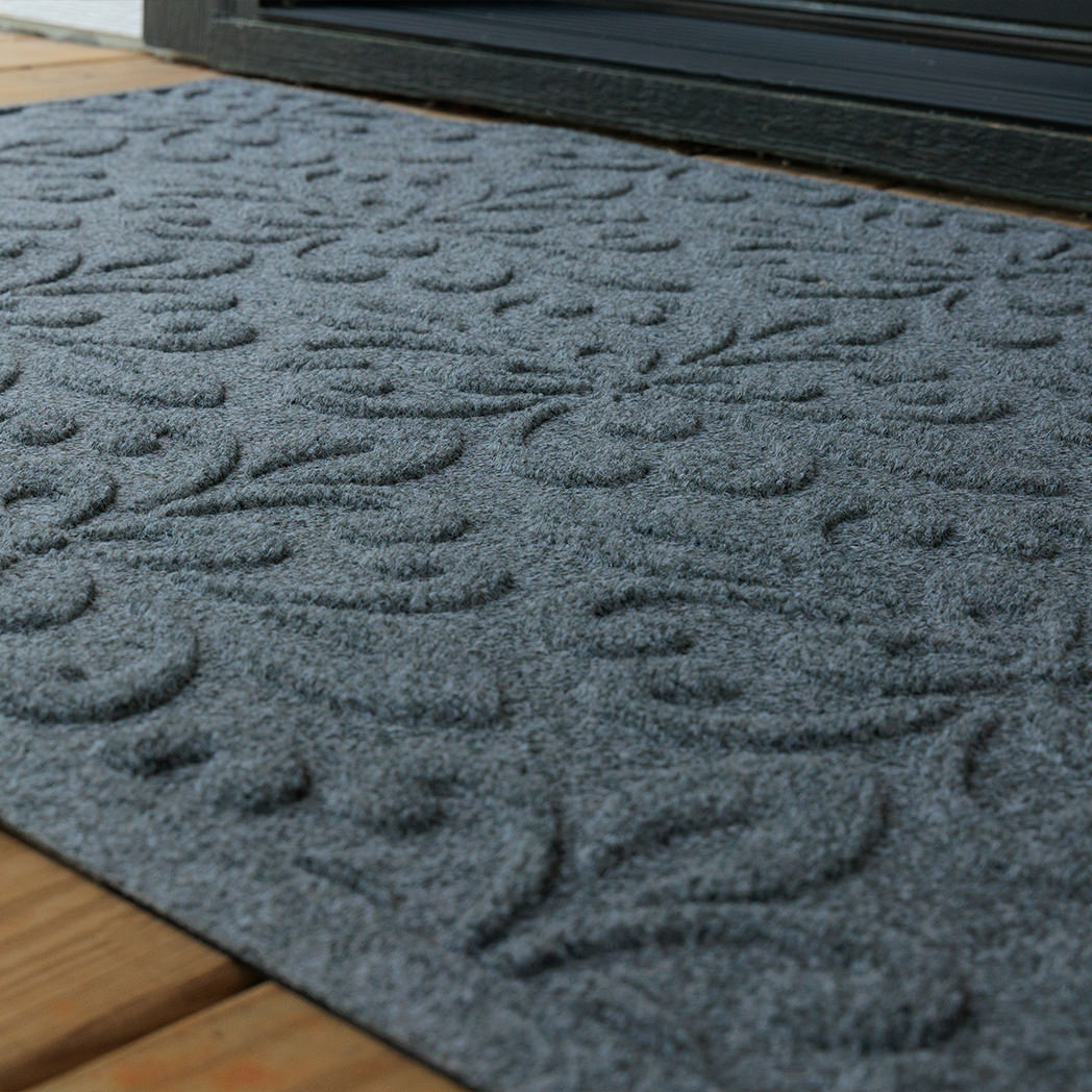 Low angled close-up of a blue-grey WaterHog Phoenix doormat on a wooden porch outside a doorway. The photo focuses on the mat’s raised floral pattern and textured surface under natural light.