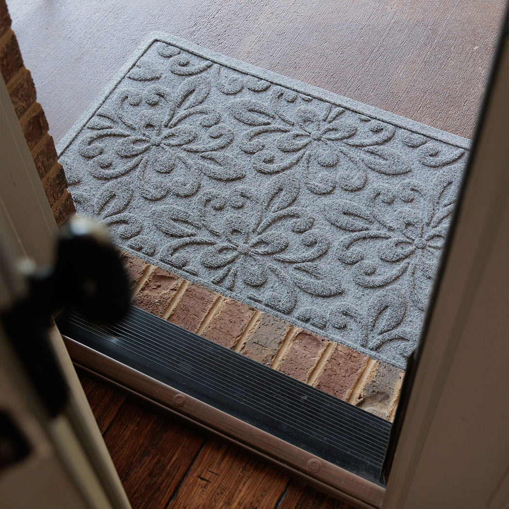 Angled view from inside a doorway looking out onto a light grey WaterHog Phoenix doormat resting on a brick threshold and concrete porch. The photo shows the mat’s raised floral pattern framed by the open door and dark wood floor inside.