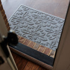 Angled view from inside a doorway looking out onto a light grey WaterHog Phoenix doormat resting on a brick threshold and concrete porch. The photo shows the mat’s raised floral pattern framed by the open door and dark wood floor inside.