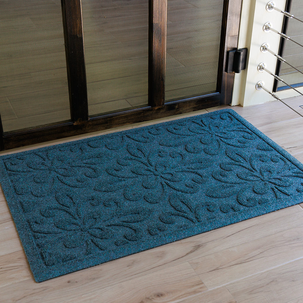 Overhead view of a blue-toned WaterHog Phoenix doormat placed just inside a dark wood-framed doorway on light wood floors. The mat’s floral-inspired raised pattern is clearly visible, showing the textured loops and recessed channels that define its surface.