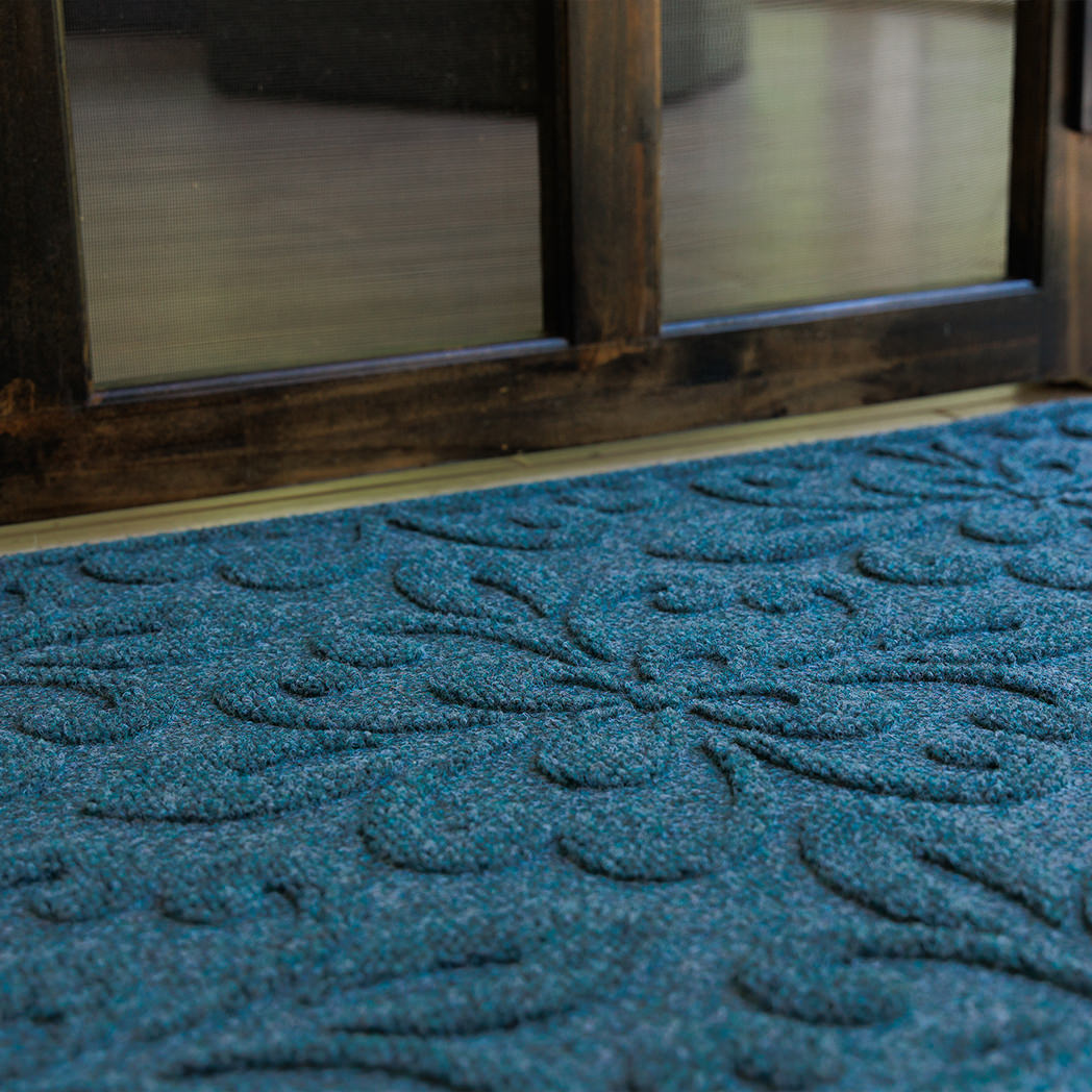 Low angled close-up of a deep blue WaterHog Phoenix doormat set just inside a dark wood-framed doorway. The photo highlights the mat’s sculpted floral pattern and textured surface against the light tan flooring.