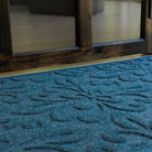 Low angled close-up of a deep blue WaterHog Phoenix doormat set just inside a dark wood-framed doorway. The photo highlights the mat’s sculpted floral pattern and textured surface against the light tan flooring.