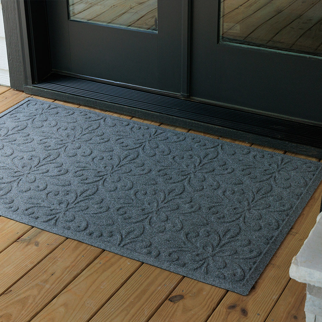 Overhead view of a large blue-grey WaterHog Phoenix doormat placed in front of black double doors on a wooden porch. The photo shows the mat’s raised floral pattern and broad coverage across the entryway.