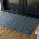 Overhead view of a large blue-grey WaterHog Phoenix doormat placed in front of black double doors on a wooden porch. The photo shows the mat’s raised floral pattern and broad coverage across the entryway.