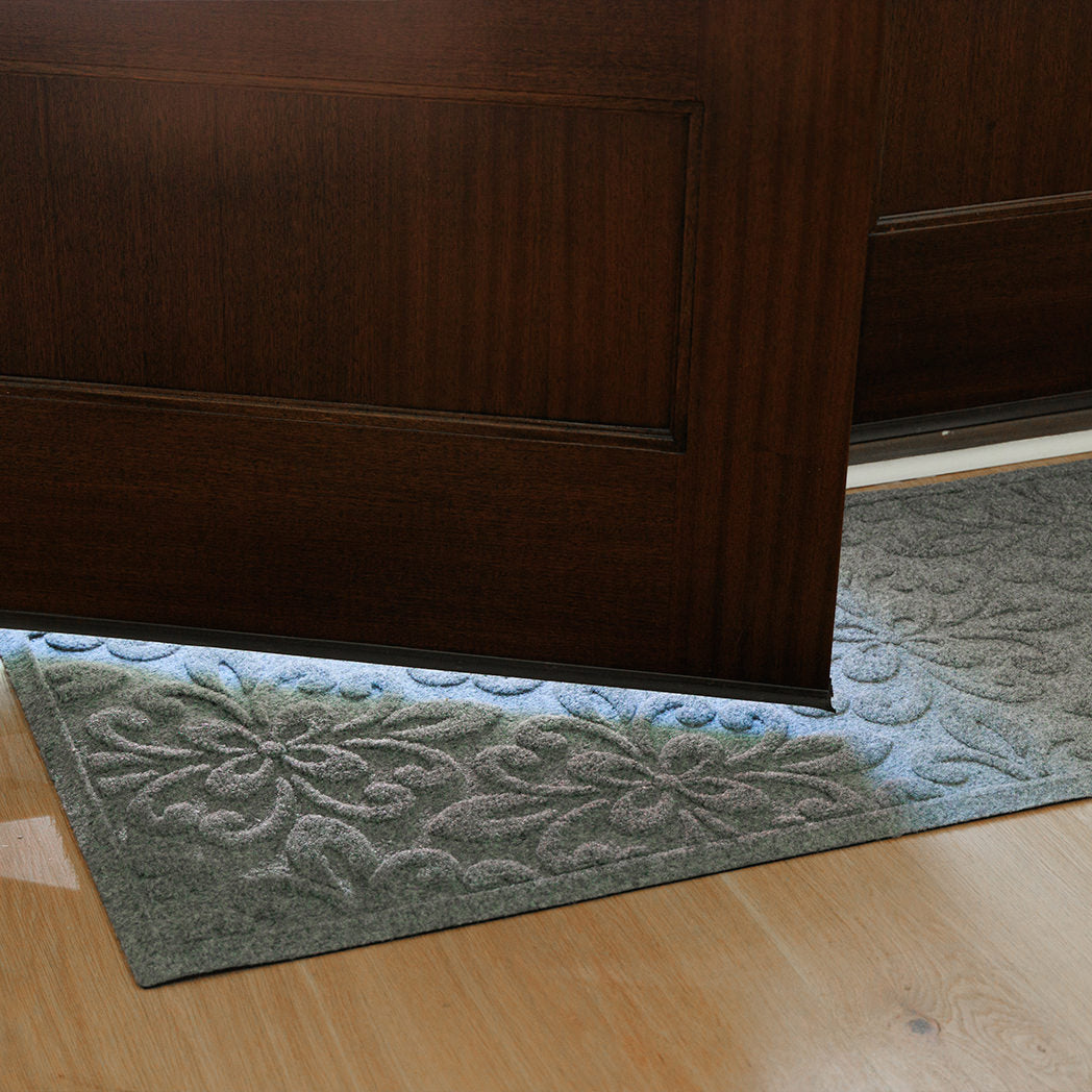 Close-up view of a grey WaterHog Phoenix runner partially covered by a dark wooden door. Sunlight passes through the gap at the bottom of the door, highlighting the mat’s raised floral pattern against the light wood floor.