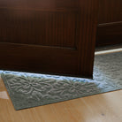 Close-up view of a grey WaterHog Phoenix runner partially covered by a dark wooden door. Sunlight passes through the gap at the bottom of the door, highlighting the mat’s raised floral pattern against the light wood floor.