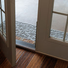 A view through an open glass door showing a grey WaterHog Phoenix runner placed outside on a concrete porch. The mat sits just beyond a brick threshold, its raised floral pattern clearly visible in the light.