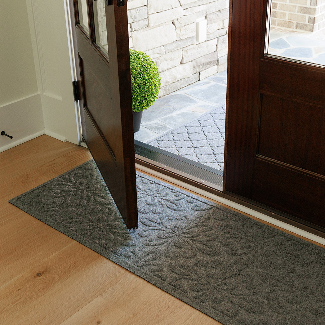 Angled view of a grey WaterHog Phoenix runner placed inside a wooden doorway on light wood flooring, with a matching grey WaterHog Cordova doormat visible just outside on a stone porch. The open door reveals the complimenting contrast between the two textured patterns.