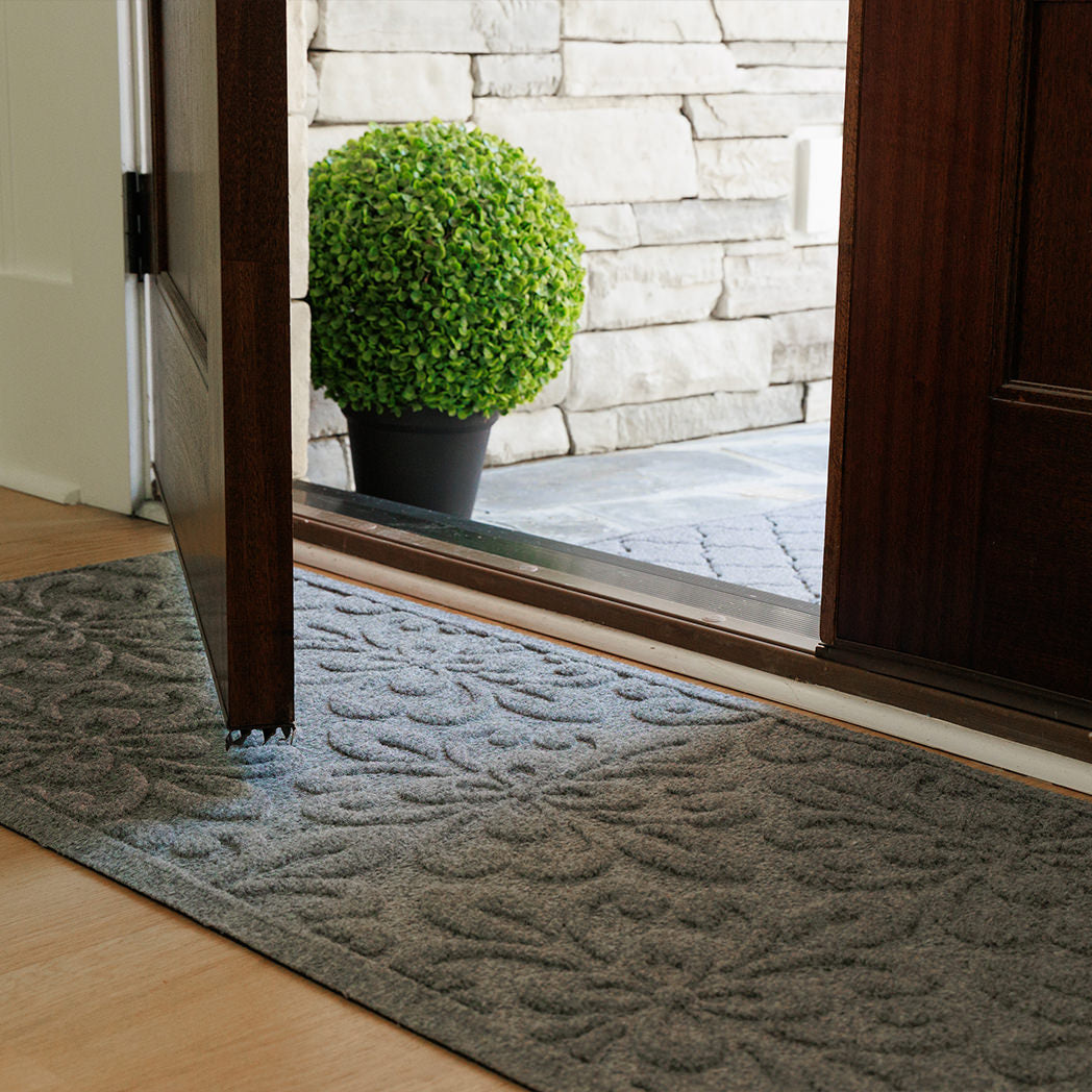 Angled view of a grey WaterHog Phoenix runner placed inside an open wooden doorway on light wood flooring. Outside, a potted green plant sits on a stone porch beside the corner of a grey WaterHog Cordova doormat.
