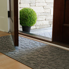 Angled view of a grey WaterHog Phoenix runner placed inside an open wooden doorway on light wood flooring. Outside, a potted green plant sits on a stone porch beside the corner of a grey WaterHog Cordova doormat.