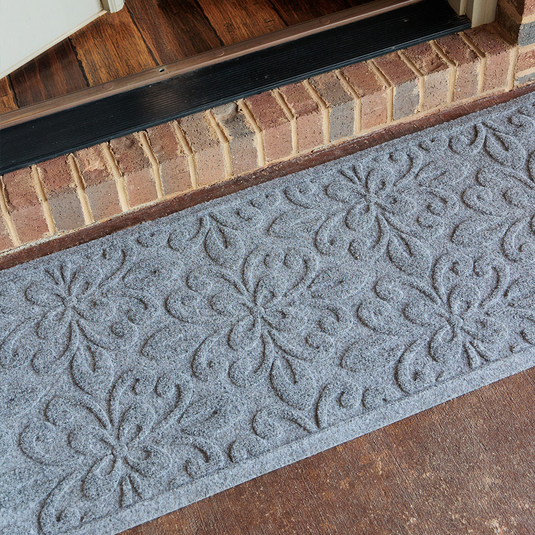 Overhead shot of a grey WaterHog Phoenix runner placed on brown-stained concrete outside the doorway of a brick house. The photo highlights the mat’s raised flowery pattern and elongated shape designed for entryway coverage.