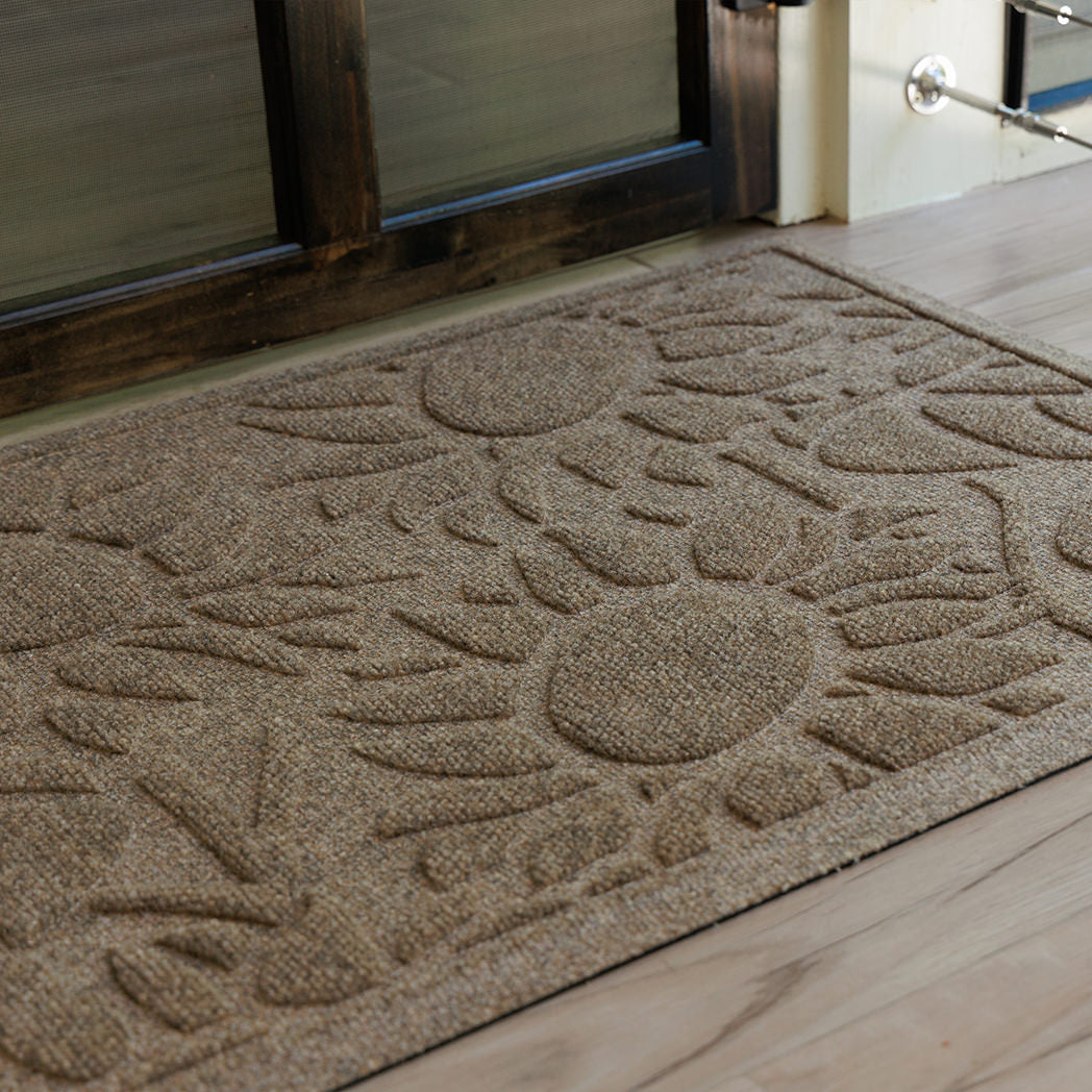 Detailed close-up of a tan WaterHog Sunshine Day mat resting on a light wood porch outside a dark-framed glass door. The shot shows the mat’s radiating sunburst design and two-tier surface, where raised ridges brush away dirt and recessed channels hold what’s left behind.