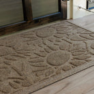 Detailed close-up of a tan WaterHog Sunshine Day mat resting on a light wood porch outside a dark-framed glass door. The shot shows the mat’s radiating sunburst design and two-tier surface, where raised ridges brush away dirt and recessed channels hold what’s left behind.