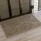 Overhead view of a tan WaterHog Sunshine Day doormat sitting just inside a dark wood-framed glass door on light wood floors. The mat’s raised sunflower pattern stands out in the soft natural light, showing the textured loops and recessed grooves of its design.