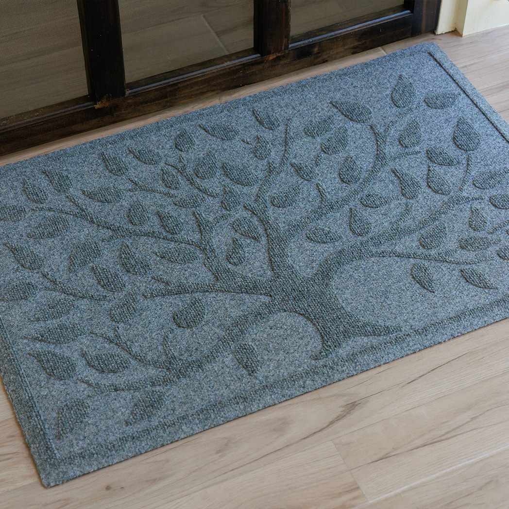 Angled close-up of a blue-grey WaterHog Tree of Life doormat sitting on a light wood porch outside a dark wood-framed glass door. The photo shows the raised tree and leaf design across the textured surface of the mat.