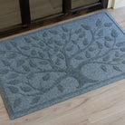 Angled close-up of a blue-grey WaterHog Tree of Life doormat sitting on a light wood porch outside a dark wood-framed glass door. The photo shows the raised tree and leaf design across the textured surface of the mat.