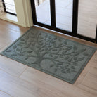 Angled overhead view of a blue-grey WaterHog Tree of Life doormat just inside a dark wood-framed glass door. The raised design shows a spreading tree with textured leaves branching across the surface, sitting on light wood flooring.