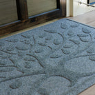 Close-up view of a blue-grey WaterHog Tree of Life doormat on a light wood porch outside a dark wood-framed glass door, displaying its two distinct levels — the top for scraping dirt and the lower for catching and holding moisture and debris.